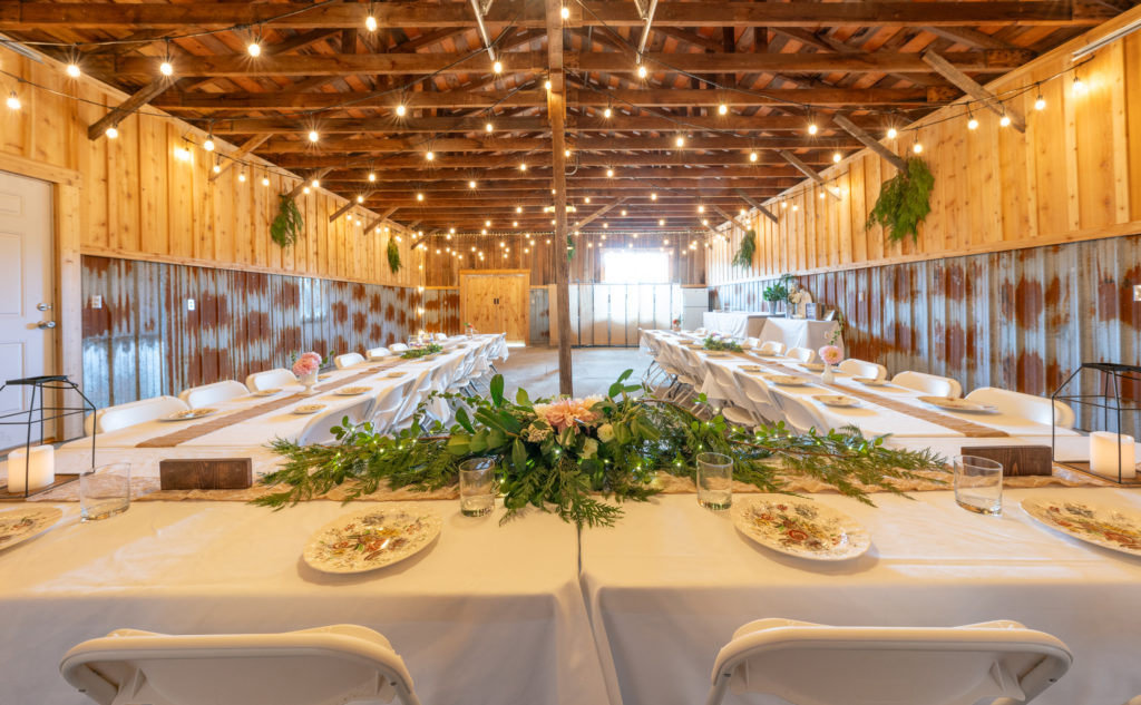 Tables set for a wedding with plates inside a barn with half the walls in rustic tin and cedar boards on the top half with open rafters and string and twinkle lights above